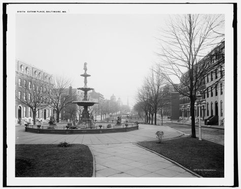 Black and white, historic photo of Eutaw Square, Baltimore, on a sunny day, with a large public fountain surrounded by high end apartment buildings and trees lining the street on both sides with long-range view toward downtown.