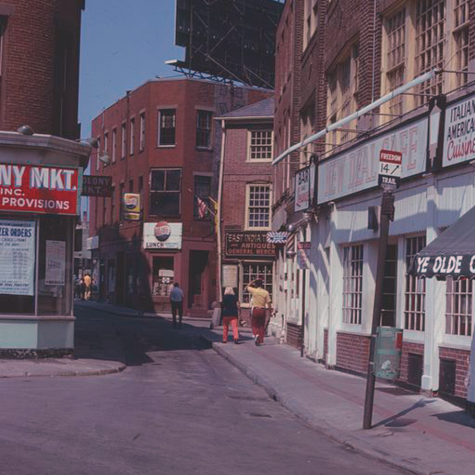A color photograph of a neighborhood street in Boston with mixed zoning. Commercial stores and markets are on the first floors; residential apartments are located above them. The street does not have any cars, but people are visible on the sidewalk in the distance.