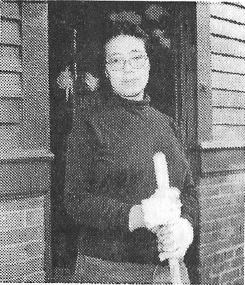 An Asian American woman stands in her doorway holding a broom, wearing work gloves and glasses.